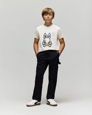 A young boy stands against a plain light gray background, wearing a white t-shirt with a cartoon bunny, Psycho Bunny KIDS RUSSELL CARPENTER PANT - KB0600001 with cargo pockets, and white sneakers. Hands in pockets, he looks at the camera.