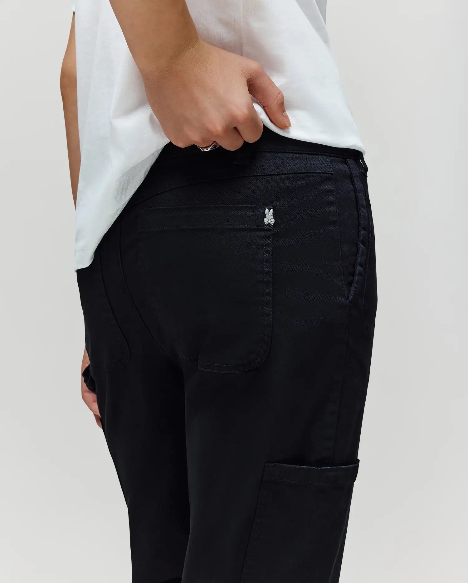 A person in a white shirt and Psycho Bunny KIDS RUSSELL CARPENTER PANT (KB0600001) stands with one hand on the waistband near the back pocket, showing a small silver butterfly emblem on the pocket.