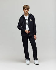 A teenage boy stands against a plain light background, wearing a black zip-up hoodie, Psycho Bunny’s white t-shirt with a large black cartoon bunny graphic, white sneakers, and KIDS RUSSELL CARPENTER PANT - KB0600001.