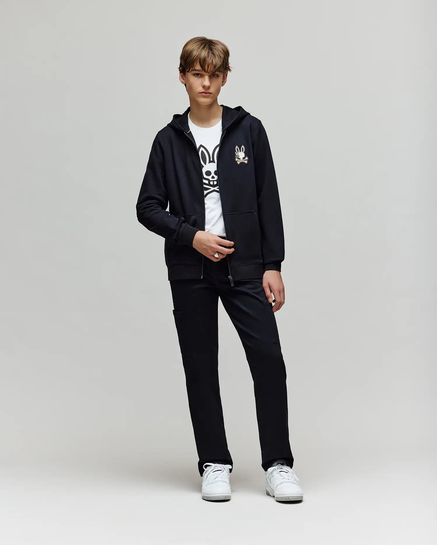 A teenage boy stands against a plain light background, wearing a black zip-up hoodie, Psycho Bunny’s white t-shirt with a large black cartoon bunny graphic, white sneakers, and KIDS RUSSELL CARPENTER PANT - KB0600001.