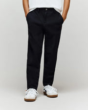 A person wears Psycho Bunny's KIDS MATHIEU COMMUTER PANT (KB0600002) featuring subtle Bunny embroidery with white sneakers, standing hands-in-pockets against a plain light background; their upper body is mostly out of frame.