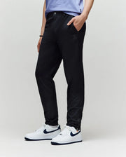 A person wears Psycho Bunny KIDS MATHIEU COMMUTER PANT (KB0600002) in black with Bunny embroidery, paired with white Nike sneakers featuring black swooshes and a light purple T-shirt, standing hands-in-pockets against a plain light background.