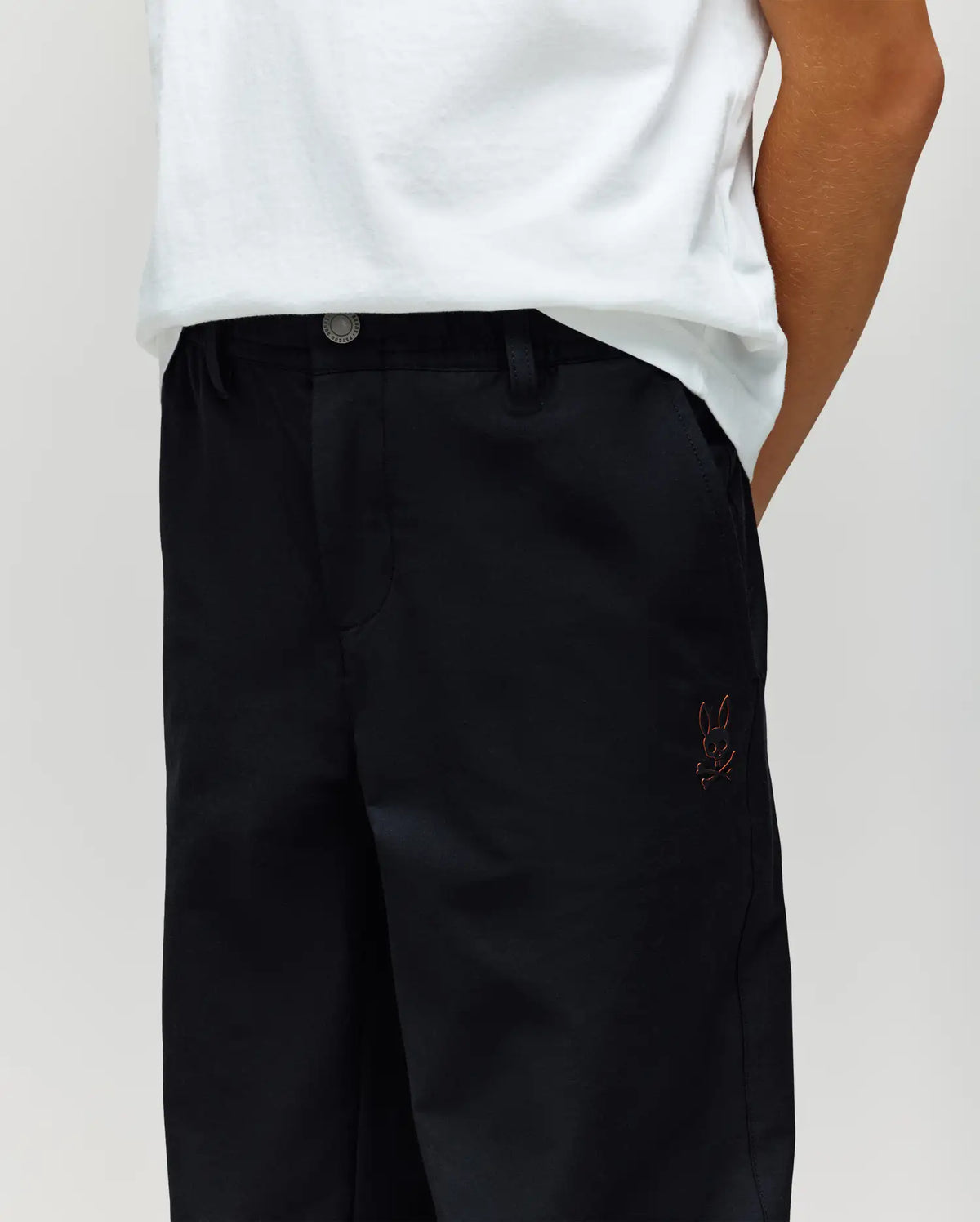 A person wears a white t-shirt tucked into high-waisted, loose-fit Psycho Bunny KIDS MATHIEU COMMUTER PANT (KB0600002) with a classic cut and small bunny embroidery on the pocket. Their face is hidden with hands behind their back.