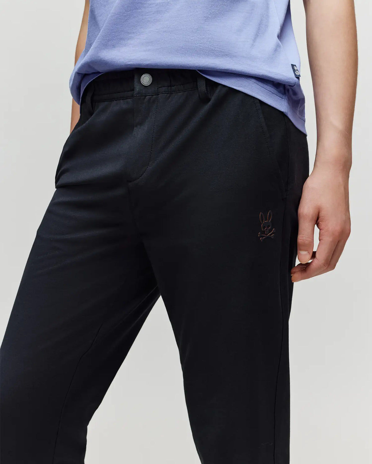 A person wears the Psycho Bunny KIDS MATHIEU COMMUTER PANT (KB0600002) featuring a small Bunny and crossbones logo on the pocket, paired with a light purple shirt, resting their hand on their thigh.