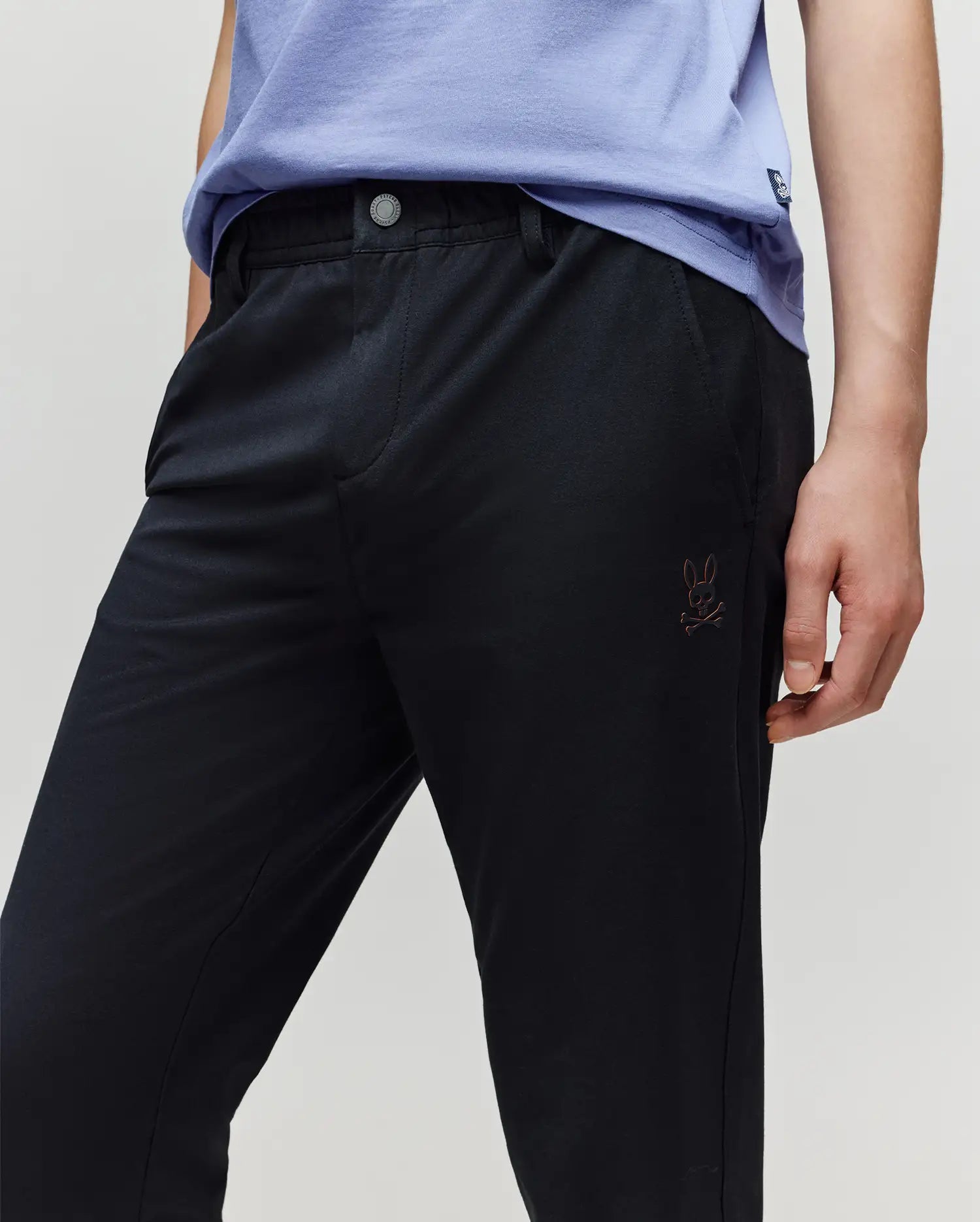 A person wears the Psycho Bunny KIDS MATHIEU COMMUTER PANT (KB0600002) featuring a small Bunny and crossbones logo on the pocket, paired with a light purple shirt, resting their hand on their thigh.
