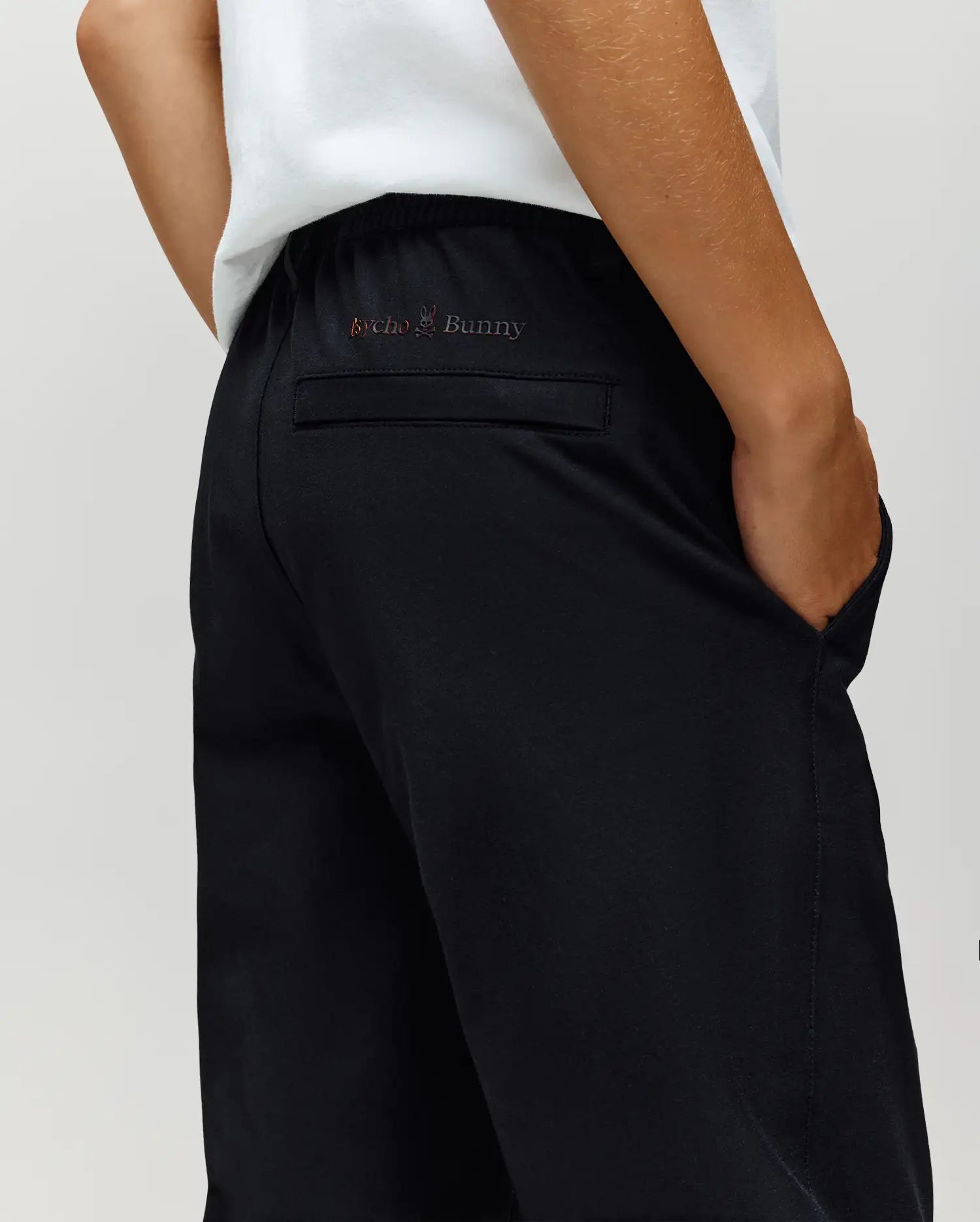 A child sports the Psycho Bunny KIDS MATHIEU COMMUTER PANT in black, featuring a classic fit and a back pocket with vibrant Bunny embroidery reading "Psycho Bunny," paired with a crisp white shirt.
