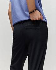 A person wears a light purple shirt and Psycho Bunny KIDS MATHIEU COMMUTER PANT (KB0600002), showing the signature Bunny embroidery above the back pocket, with one hand in their pocket and a black bracelet.
