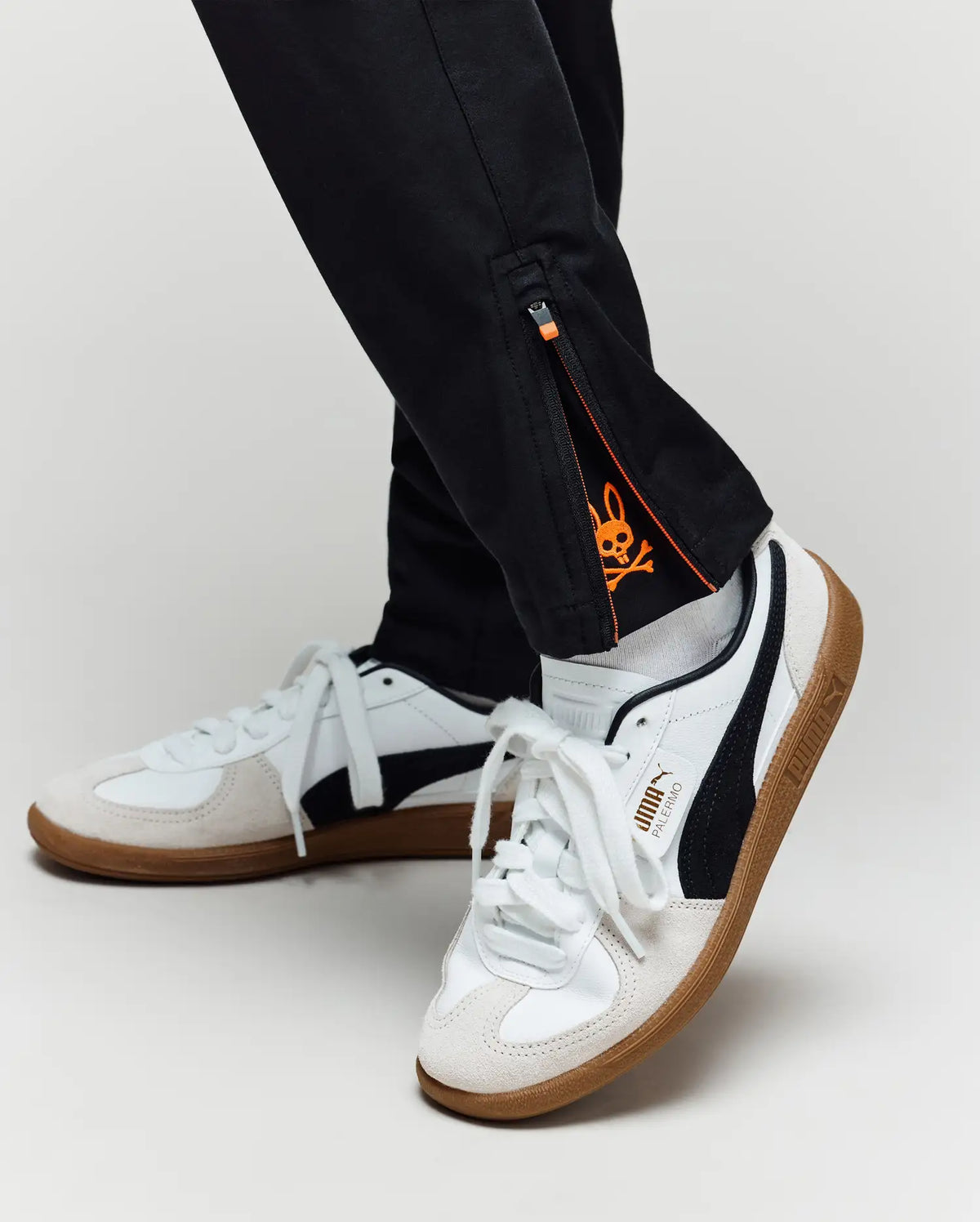 A person wears Psycho Bunny KIDS MATHIEU COMMUTER PANT (KB0600002) featuring a small orange crown logo on the inner ankle, styled with white sneakers with black stripes and gum soles.