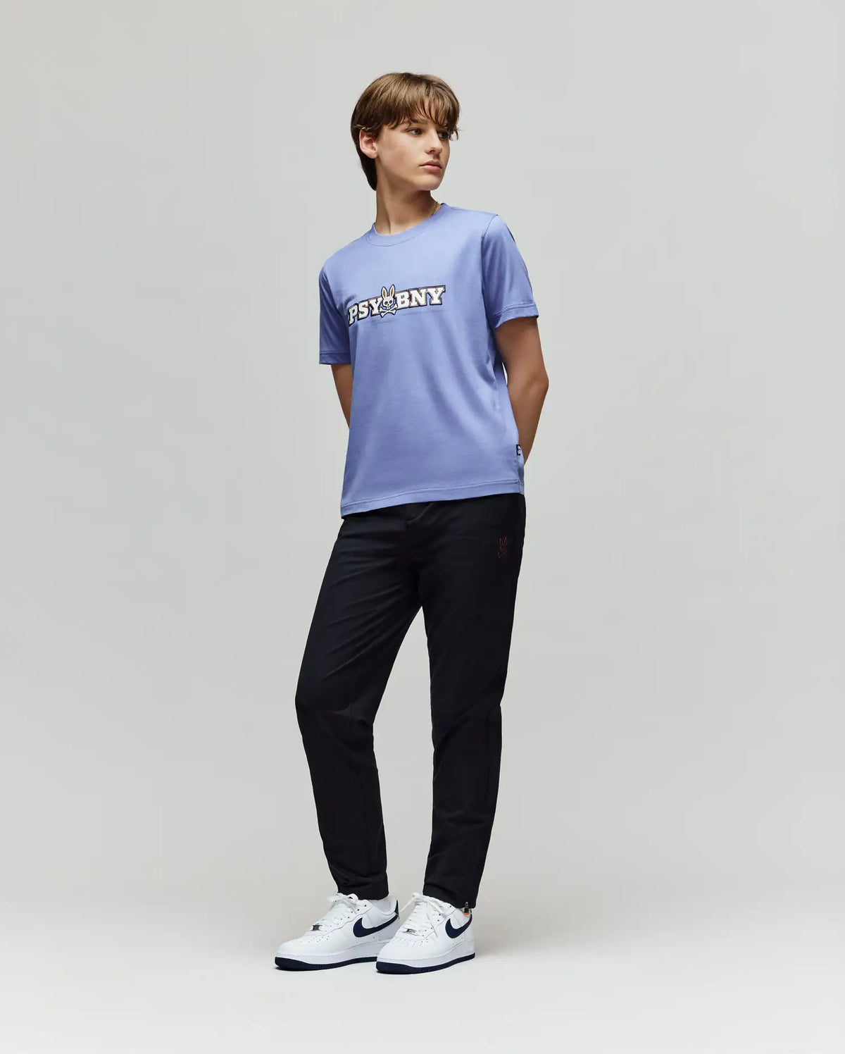 A person stands against a plain light background, wearing a classic-fit light purple T-shirt with "PSYCHNY" text, KIDS MATHIEU COMMUTER PANT - KB0600002 by Psycho Bunny, and white Nike sneakers with black swooshes.