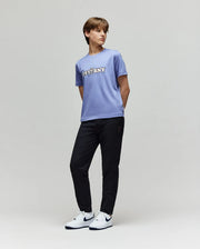 A person stands against a plain light background, wearing a classic-fit light purple T-shirt with "PSYCHNY" text, KIDS MATHIEU COMMUTER PANT - KB0600002 by Psycho Bunny, and white Nike sneakers with black swooshes.