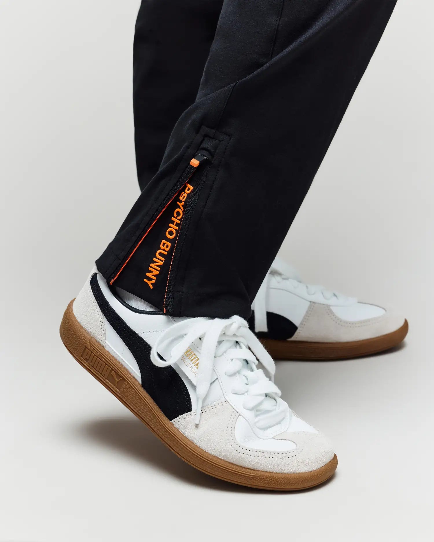 A person stands against a plain light background, wearing Psycho Bunny's KIDS MATHIEU COMMUTER PANT (KB0600002) in classic black with Bunny embroidery and an orange label, paired with white Puma sneakers with black stripes and gum soles.