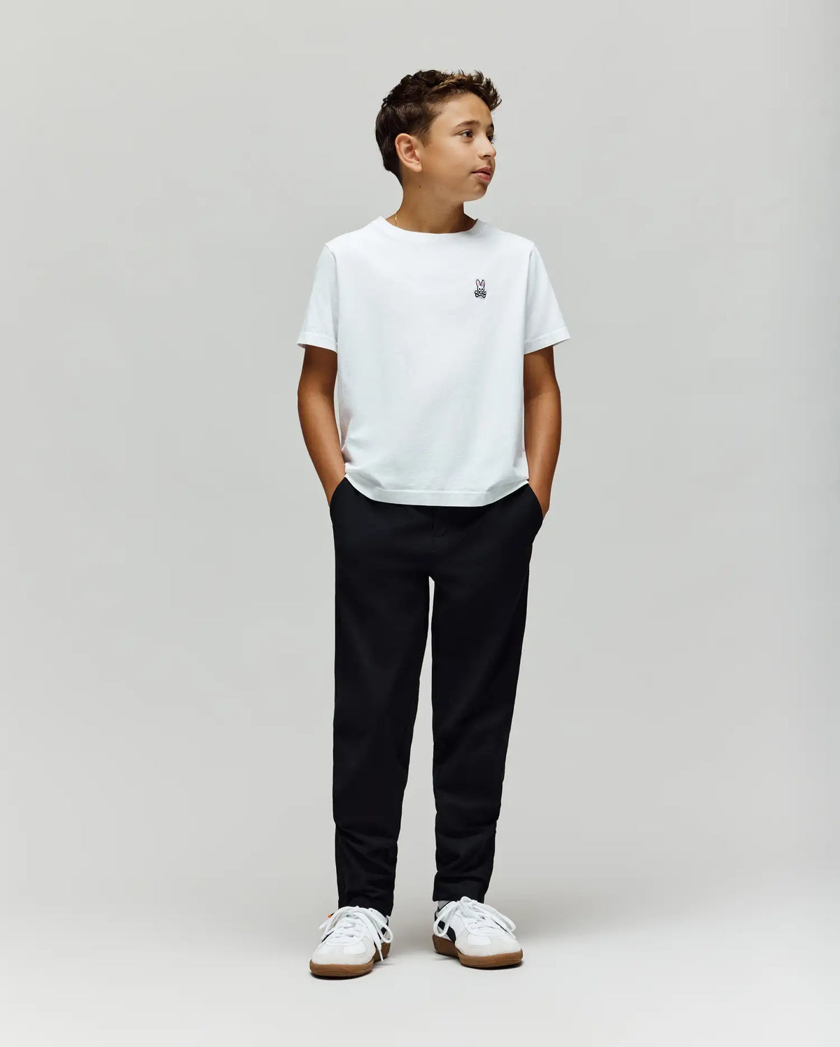 A boy stands against a plain light gray background, wearing a white t-shirt, white sneakers, and Psycho Bunny's KIDS MATHIEU COMMUTER PANT (KB0600002). His hands are in his pockets as he looks to the side.