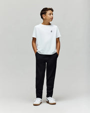A boy stands against a plain light gray background, wearing a white t-shirt, white sneakers, and Psycho Bunny's KIDS MATHIEU COMMUTER PANT (KB0600002). His hands are in his pockets as he looks to the side.