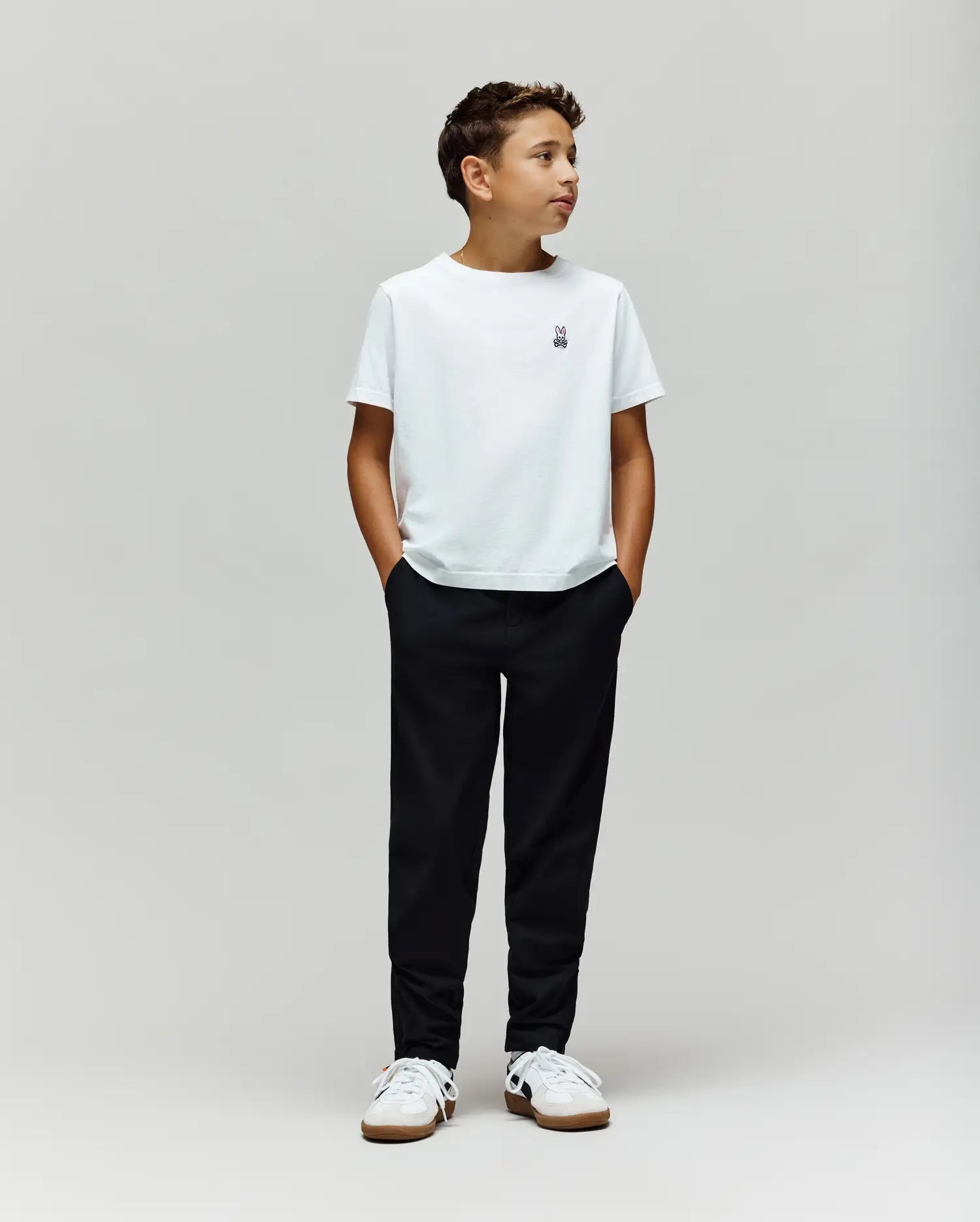 A boy stands against a plain light gray background, wearing a white t-shirt, white sneakers, and Psycho Bunny's KIDS MATHIEU COMMUTER PANT (KB0600002). His hands are in his pockets as he looks to the side.