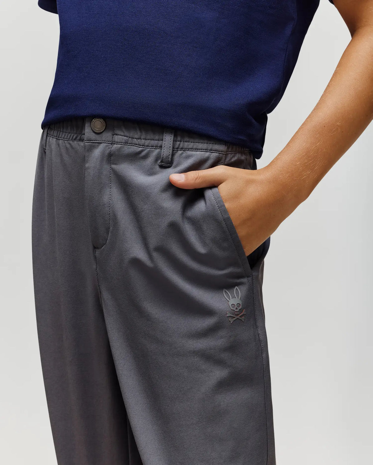 A person wearing a navy blue shirt and Psycho Bunny KIDS MATHIEU COMMUTER PANT - KB0600002 in classic fit gray, featuring bunny embroidery on the pocket, stands with their right hand in the pocket. Only torso and upper legs are shown.