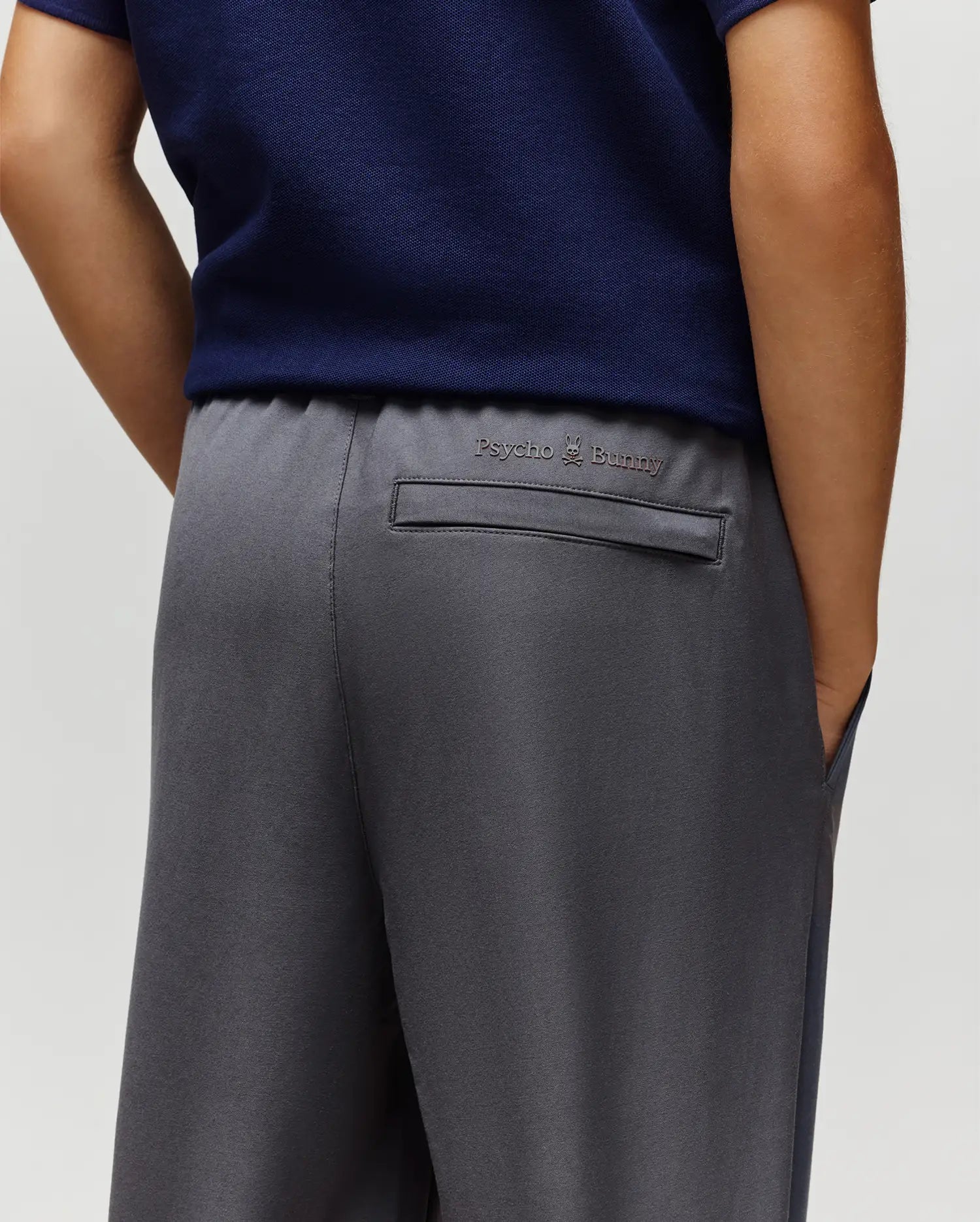 A person in a navy shirt and Psycho Bunny KIDS MATHIEU COMMUTER PANT (KB0600002) in gray, with one hand in their pocket. The trousers’ back pocket is detailed with the signature Bunny embroidery above it.