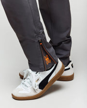 A close-up of a person wearing white Puma sneakers with black stripes and gum soles, paired with Psycho Bunny's KIDS MATHIEU COMMUTER PANT (KB0600002), classic fit with zippered cuffs and an orange graphic detail by the ankle.