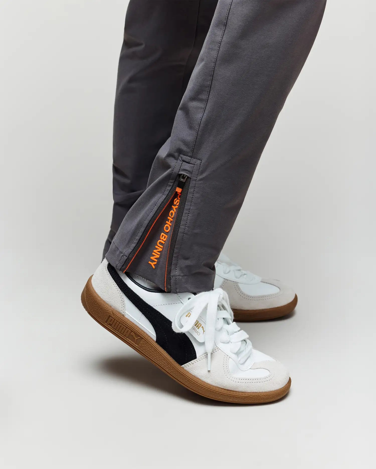 Someone wearing Psycho Bunny KIDS MATHIEU COMMUTER PANT (KB0600002) in gray, featuring orange and black zipper detail by the ankle, paired with white Puma sneakers with black accents and gum soles.