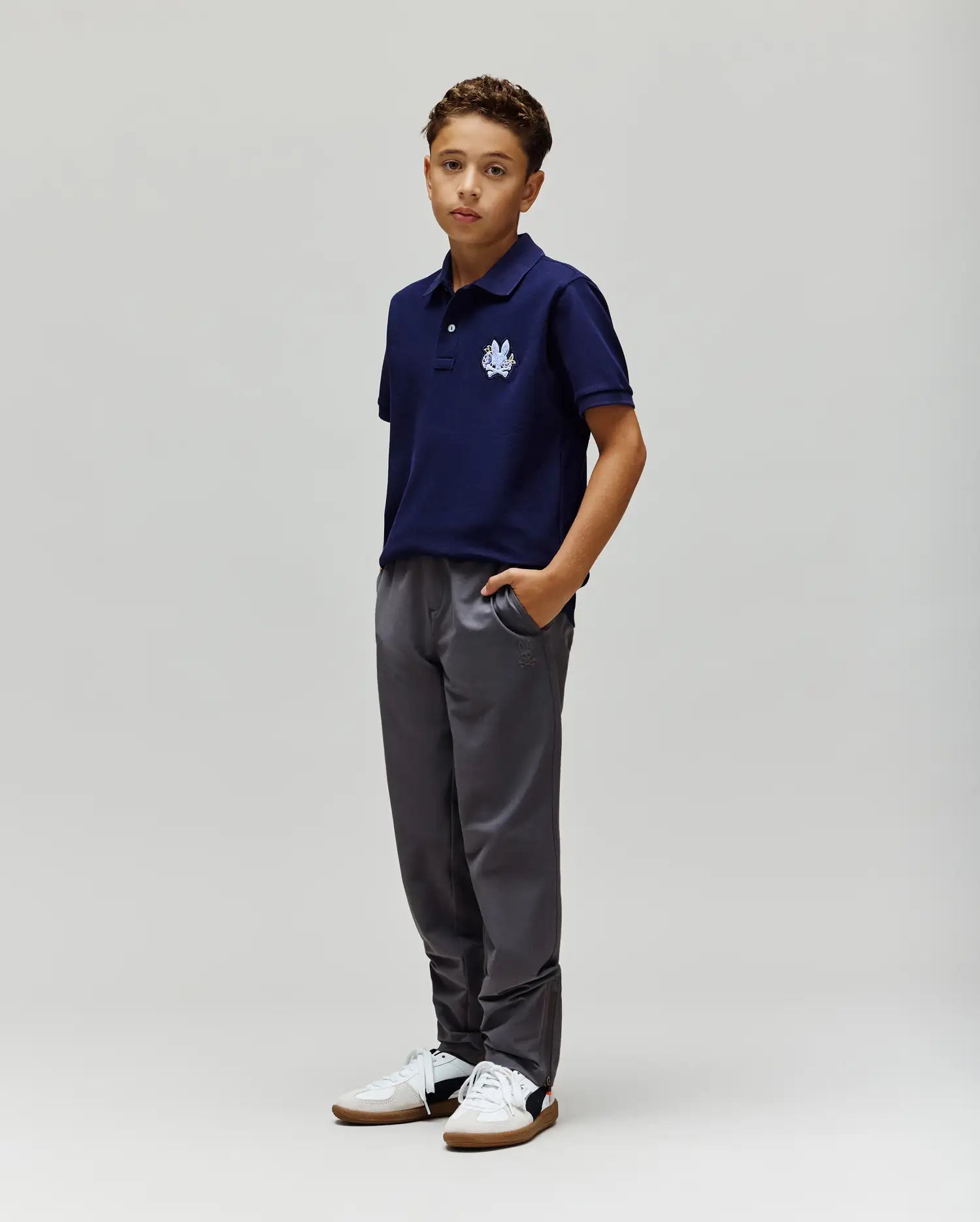 A young boy with short curly hair stands against a light gray background, wearing Psycho Bunny’s navy polo with white bunny logo, KIDS MATHIEU COMMUTER PANT - KB0600002 in gray, and white sneakers, hands in his pockets.