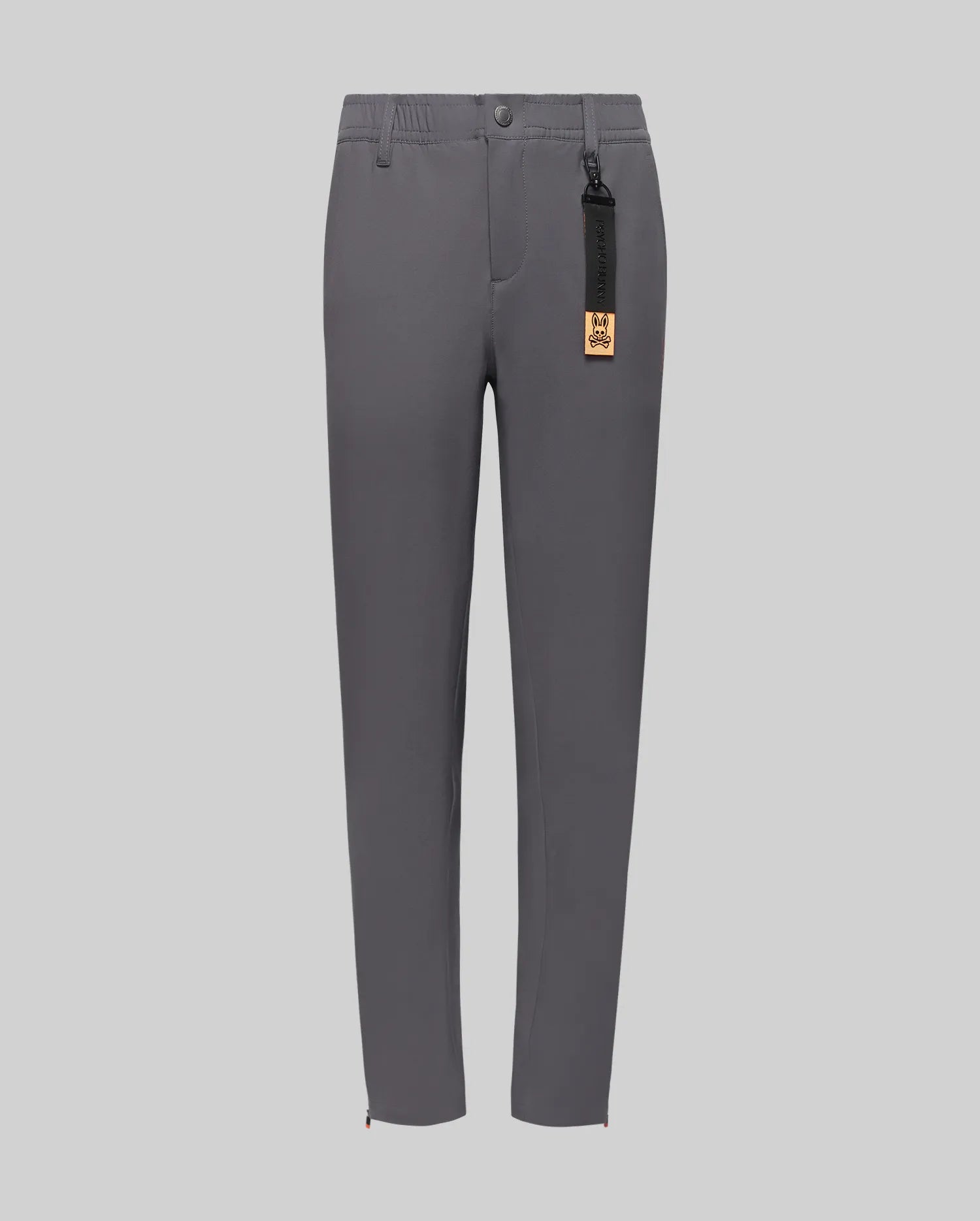 The Psycho Bunny KIDS MATHIEU COMMUTER PANT (KB0600002) is a slim-fit gray pair featuring button and zip closure, side pockets, and a black tag with gold label on the belt loop, shown against a light gray background.