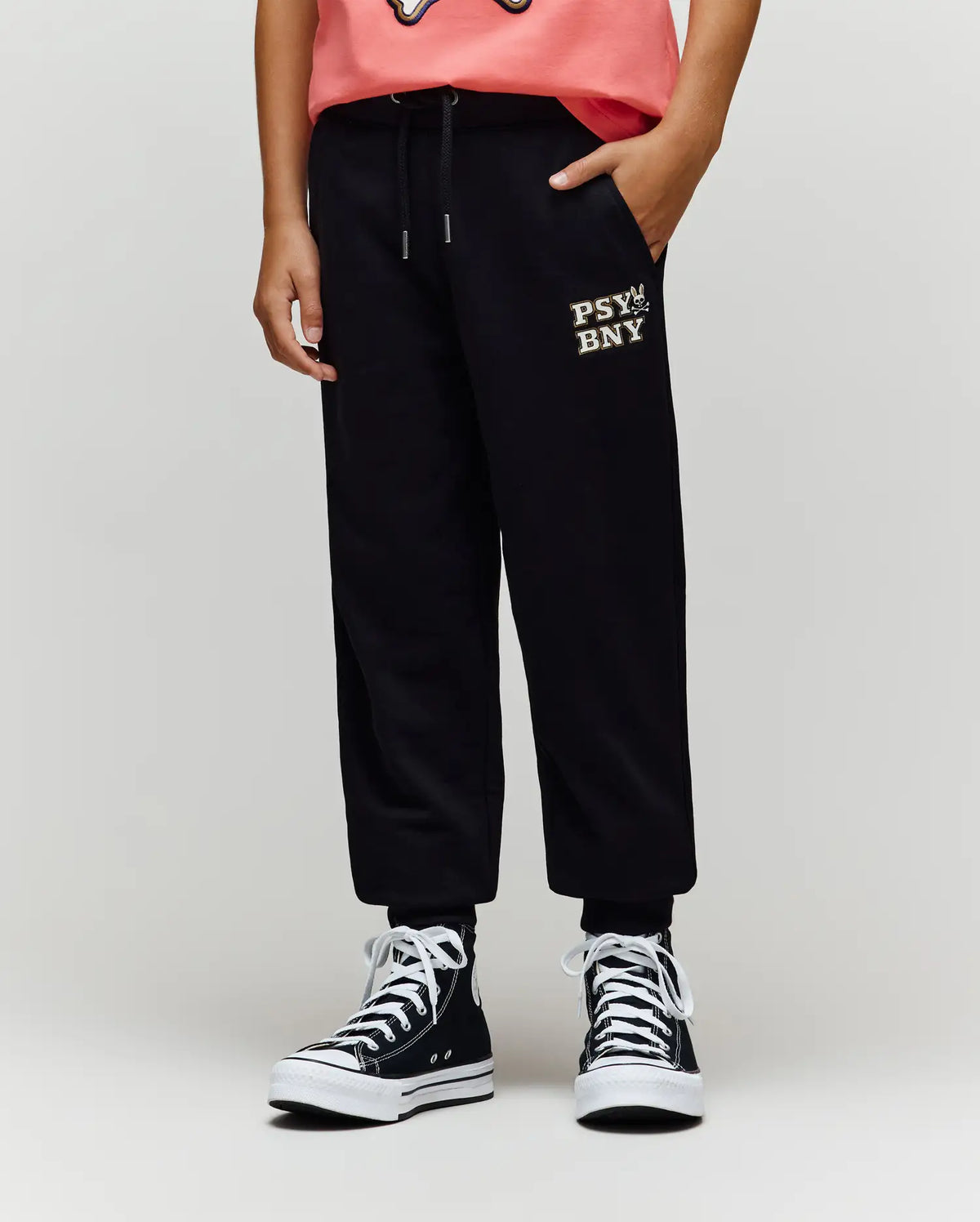 A person wears Psycho Bunny KIDS WADE SWEATPANT (KB0800002) in black, featuring an embroidered wordmark, paired with a pink shirt and black high-top sneakers. Only the lower half is visible, highlighting classic athletic style.