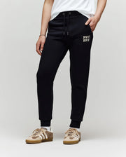 A person wearing Psycho Bunny KIDS WADE SWEATPANT - KB0800002 in black, featuring an embroidered "PSY NYC" wordmark on the left thigh, paired with a white t-shirt and beige sneakers, stands against a plain light background.
