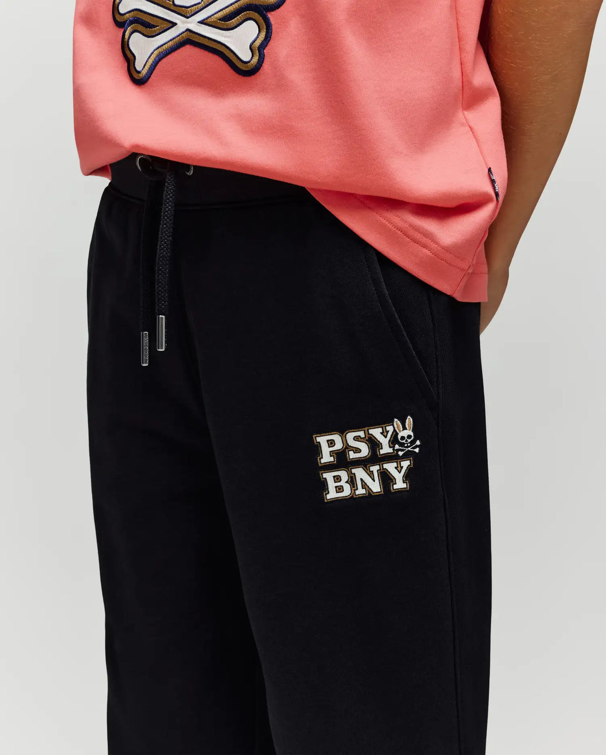 A person wearing a pink shirt with a white skull and crossbones and Psycho Bunny's KIDS WADE SWEATPANT (KB0800002), black pants featuring an embroidered "PSY BNY" wordmark and bunny logo on the upper left thigh for a classic athletic look.