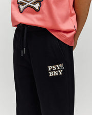 A person wearing a pink shirt with a white skull and crossbones and Psycho Bunny's KIDS WADE SWEATPANT (KB0800002), black pants featuring an embroidered "PSY BNY" wordmark and bunny logo on the upper left thigh for a classic athletic look.