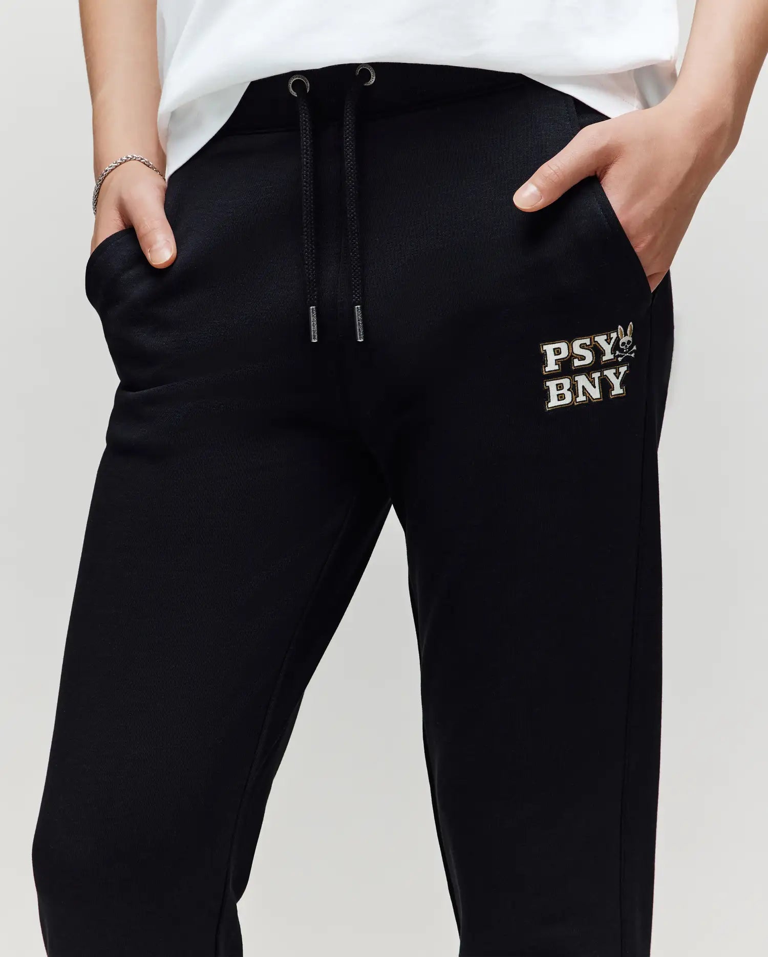 A person wears Psycho Bunny KIDS WADE SWEATPANT - KB0800002, black joggers in a premium cotton blend with "PSY BNY" and a graphic on the left thigh, hands in pockets, and a white shirt partially visible.