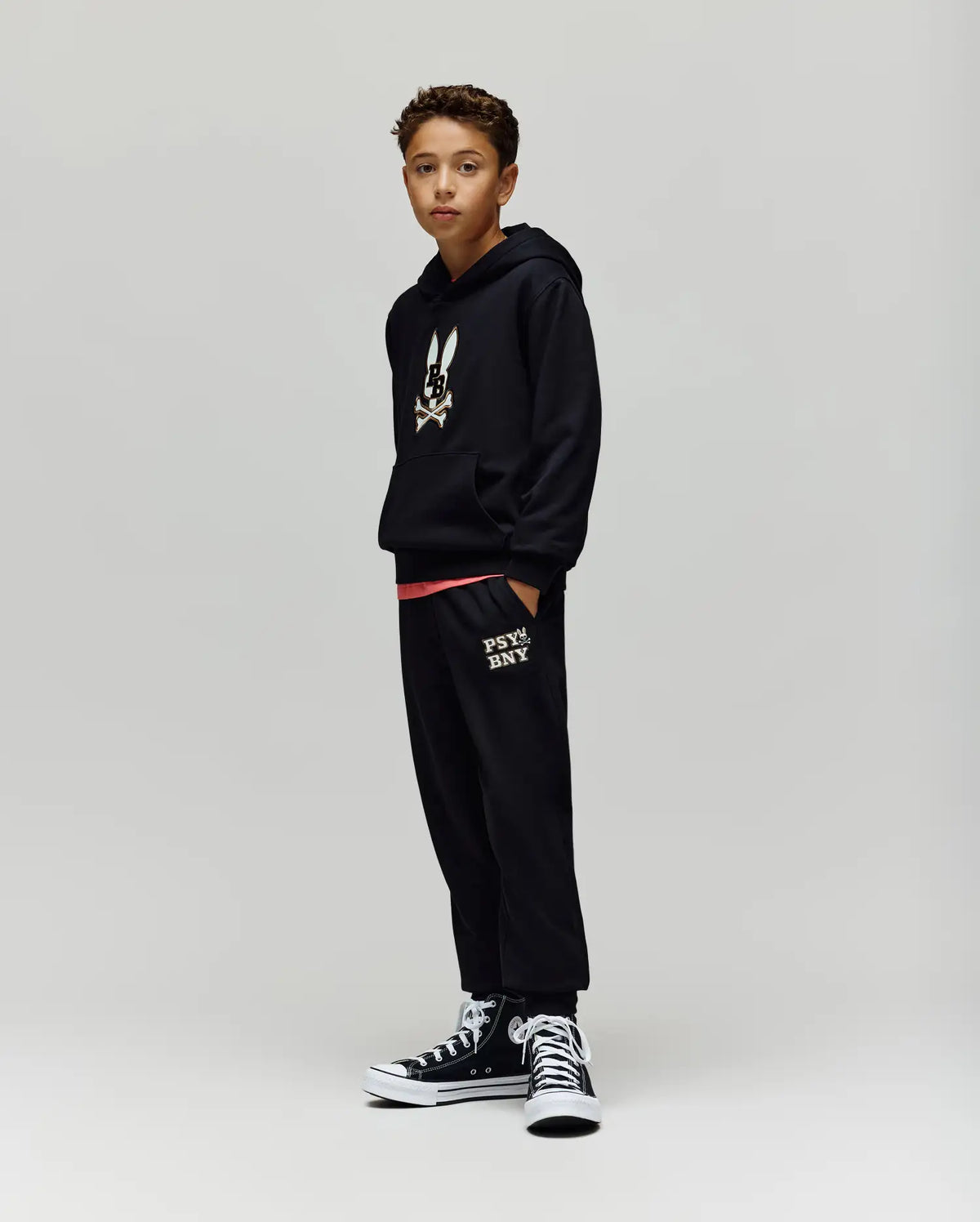 A boy stands against a plain background wearing Psycho Bunny KIDS WADE SWEATPANT (KB0800002) and a black hoodie with a cartoon bunny graphic, paired with black high-top sneakers. His hands are in his pockets as he looks at the camera.