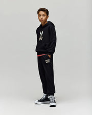 A boy stands against a plain background wearing Psycho Bunny KIDS WADE SWEATPANT (KB0800002) and a black hoodie with a cartoon bunny graphic, paired with black high-top sneakers. His hands are in his pockets as he looks at the camera.