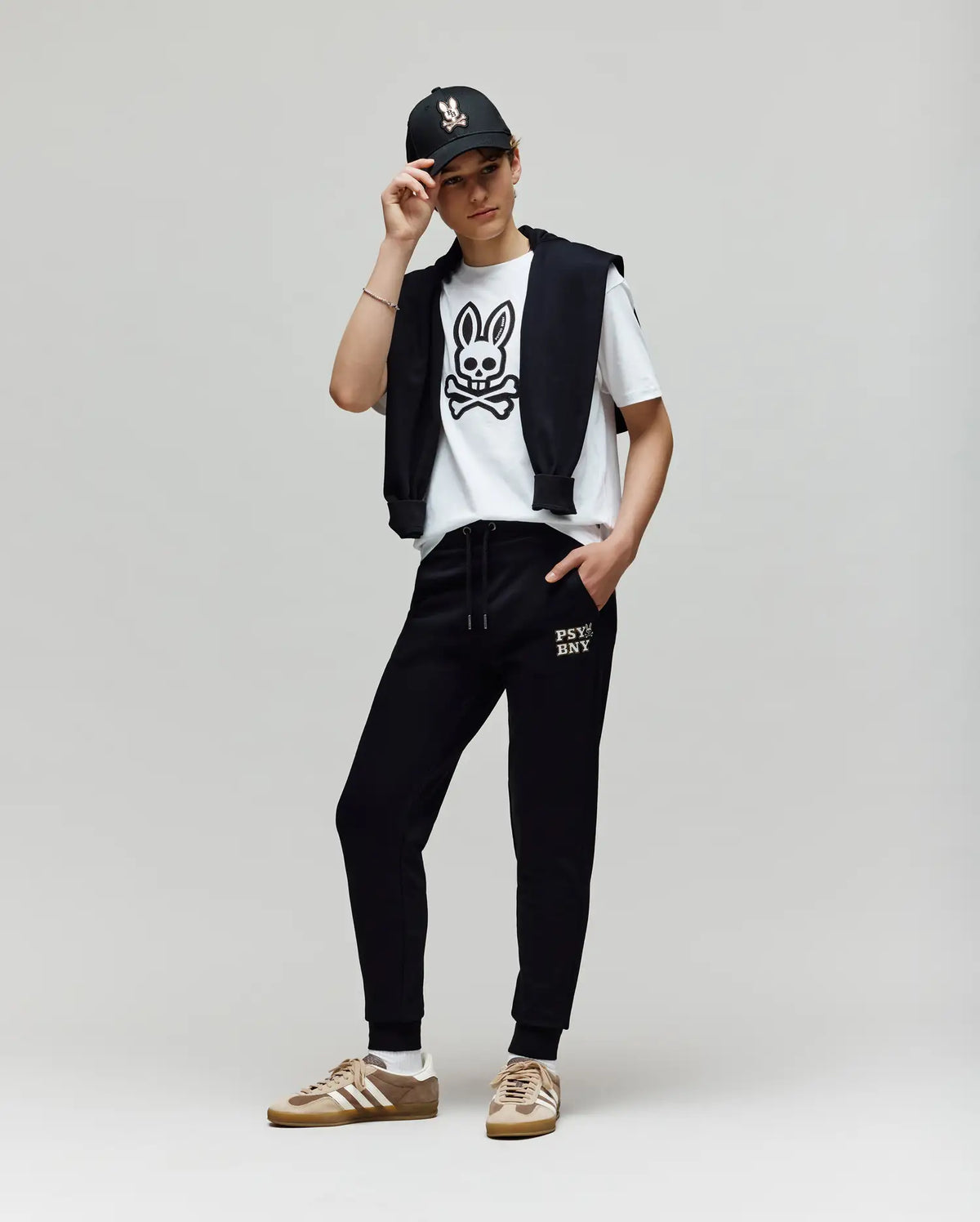 A person stands against a plain background wearing a black cap, premium cotton blend white T-shirt with a bunny skull graphic, black jacket over their shoulders, Psycho Bunny KIDS WADE SWEATPANT (KB0800002), and tan sneakers with white accents.
