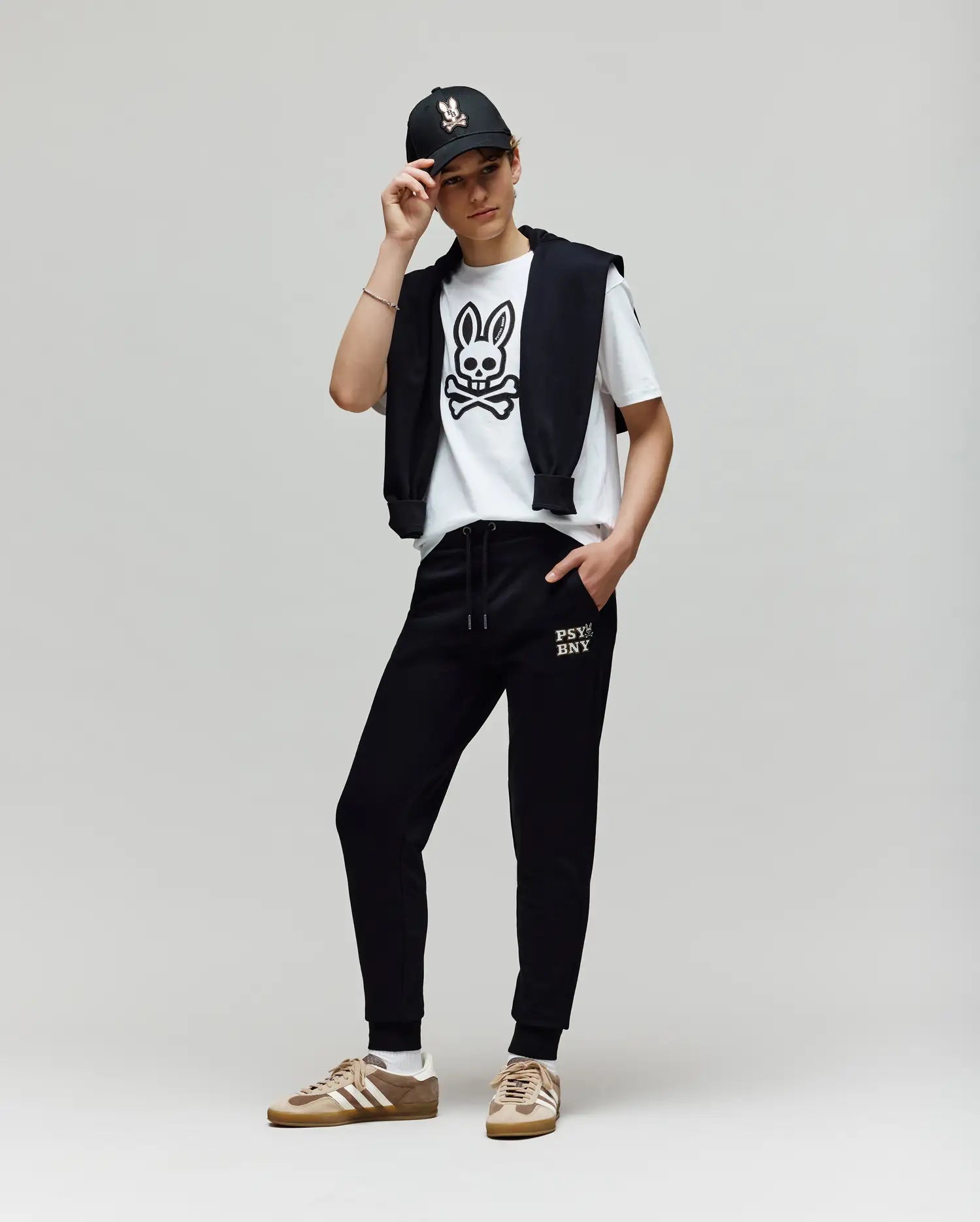 A person stands against a plain background wearing a black cap, premium cotton blend white T-shirt with a bunny skull graphic, black jacket over their shoulders, Psycho Bunny KIDS WADE SWEATPANT (KB0800002), and tan sneakers with white accents.