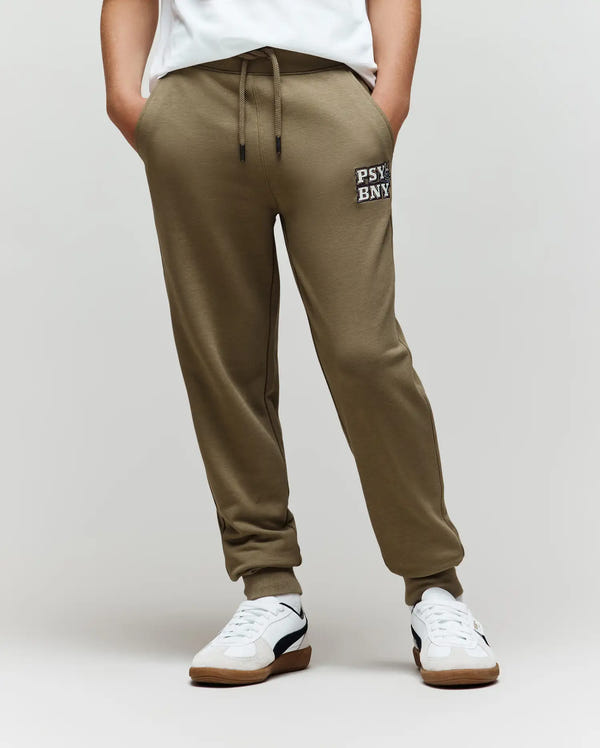 A person wearing olive green Psycho Bunny KIDS WADE SWEATPANT (KB0800002) with "PSY-BNY" on the left thigh, a white shirt, and white sneakers with black accents stands hands-in-pockets against a plain background.