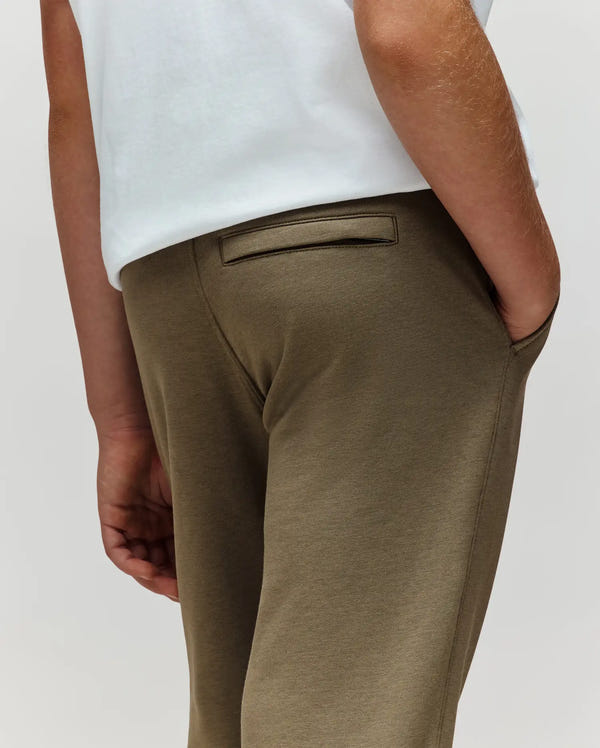 A child models Psycho Bunny’s KIDS WADE SWEATPANT (KB0800002) in olive green, paired with a white premium cotton-blend shirt, posing waist-down with one hand in their pocket against a plain background.