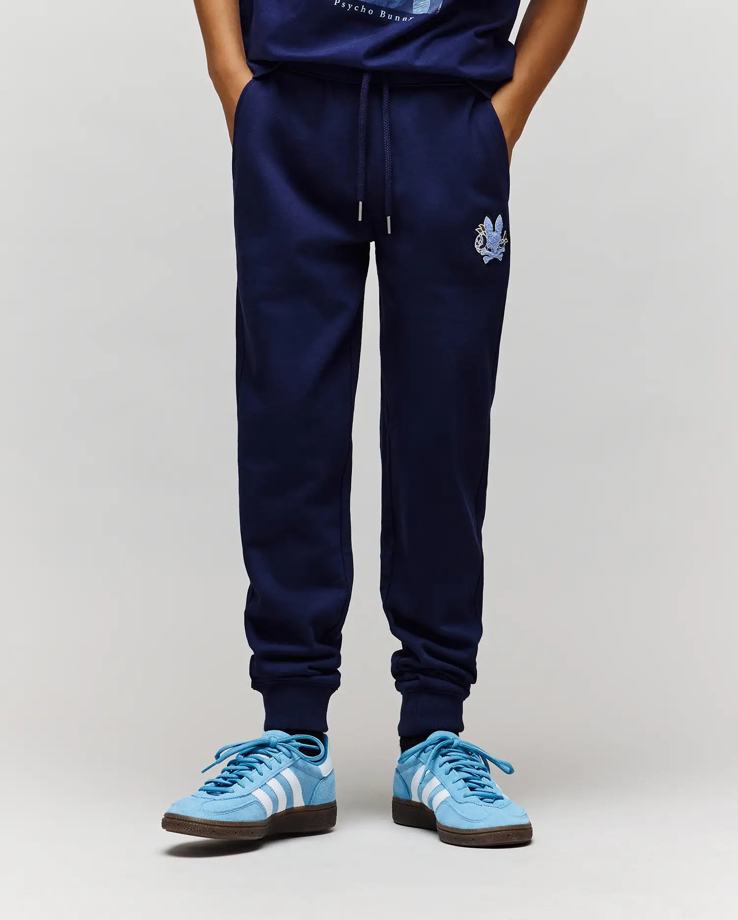 A person wears Psycho Bunny KIDS AUGUSTE JOGGER - KB0800003 in navy blue (100% cotton, small embroidered crest on left thigh) with light blue Adidas sneakers. Only the lower body is shown against a plain light grey background.