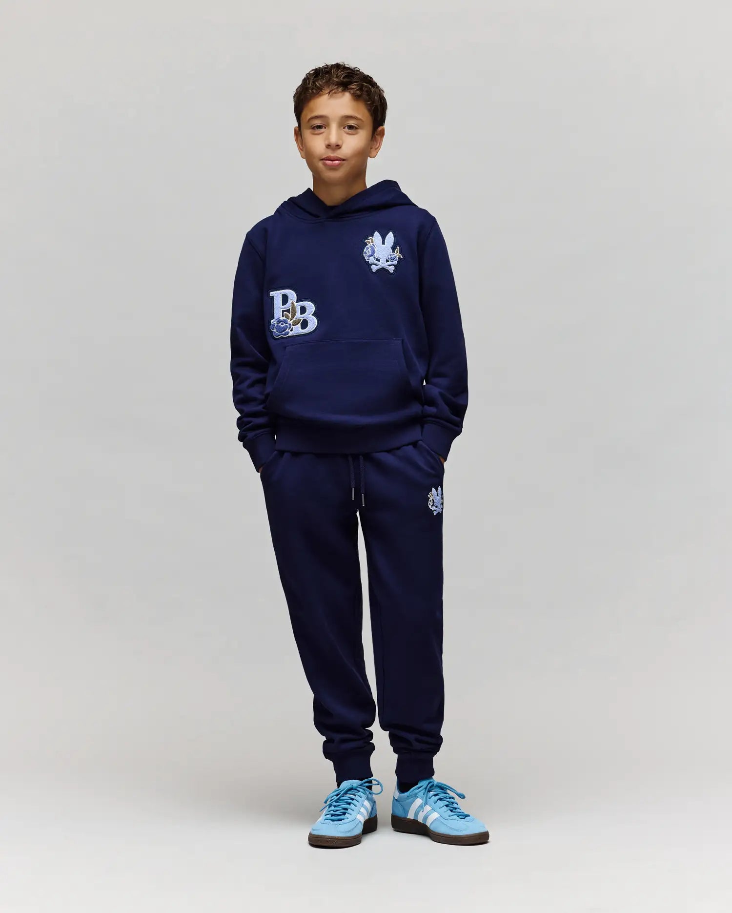 A boy stands against a plain background, wearing a navy blue hoodie, Psycho Bunny KIDS AUGUSTE JOGGER - KB0800003 with chenille patches, and light blue Adidas sneakers. His hands are in his pockets as he looks forward with a slight smile.