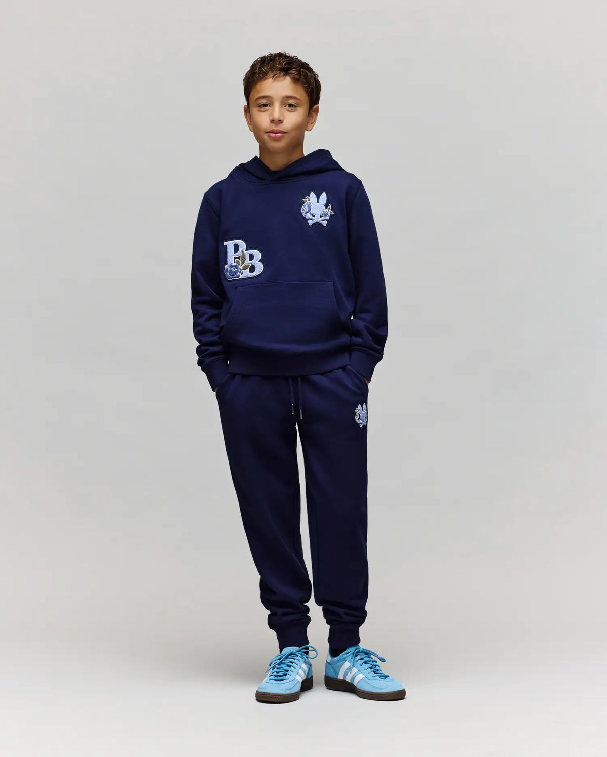 A boy models the Psycho Bunny KIDS AUGUSTE HOODIE (KT0500019), a navy cotton fleece with chenille floral accents, paired with matching joggers and light blue Adidas sneakers. He stands against a plain background, hands in pockets, smiling at the camera.