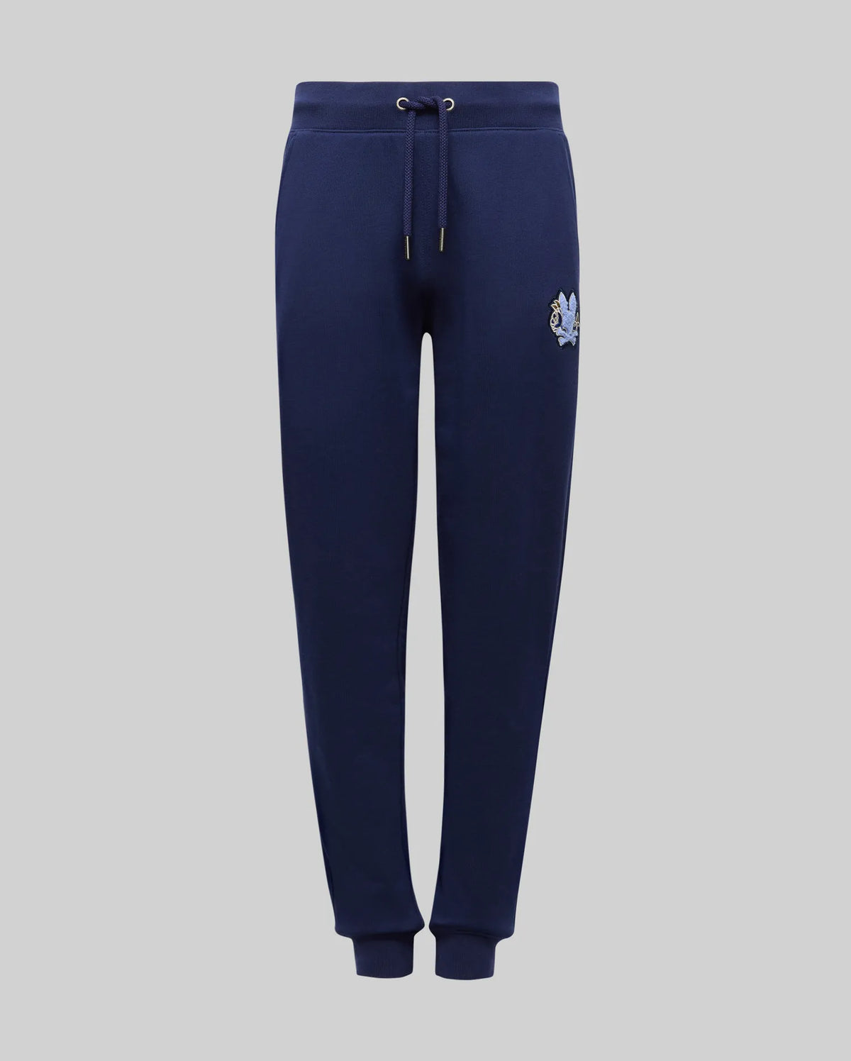 The KIDS AUGUSTE JOGGER - KB0800003 by Psycho Bunny is a navy blue 100% cotton jogger with an elastic waistband, drawstring, ribbed cuffs, and a small embroidered crest on the left thigh, shown against a plain grey background.