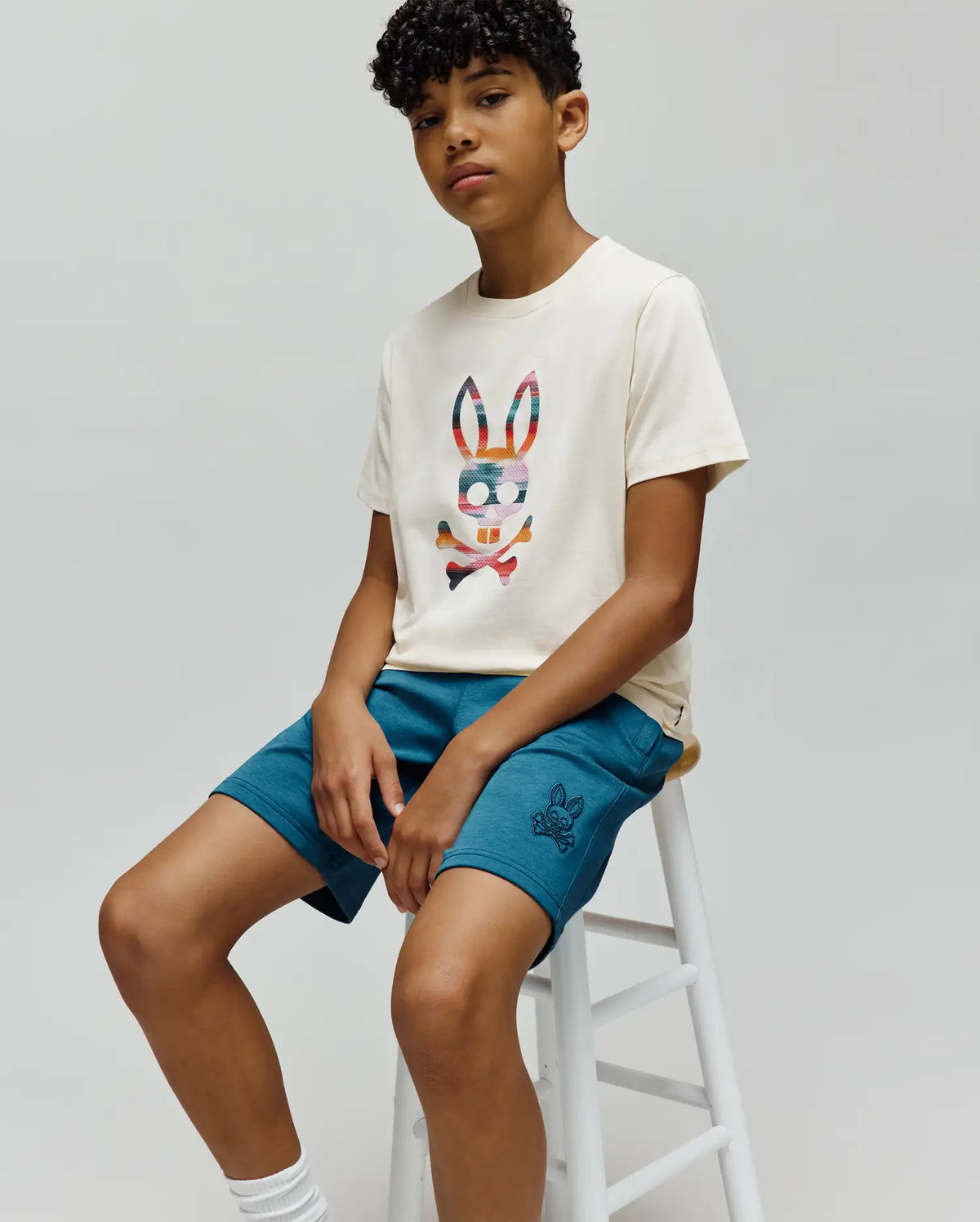 Cream jersey t-shirt with a large, colorful plaid Psycho Bunny skull graphic on the chest, paired with relaxed fit Teal KIDS BARRY MODAL SHORT featuring a small embroidered Psycho Bunny logo on the left leg.