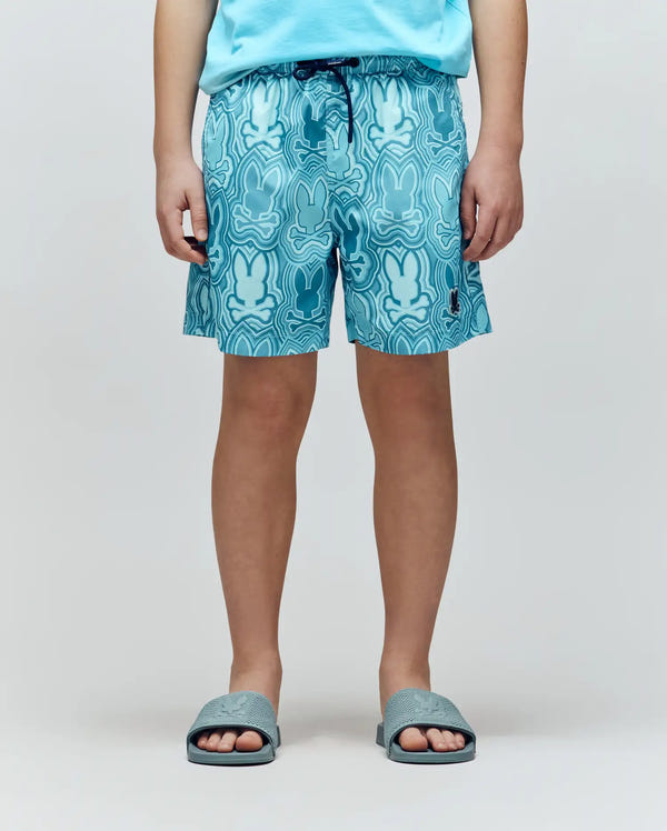 A child faces forward, wearing a blue T-shirt and Psycho Bunny KIDS CHUCK ALL OVER PRINT LIGHTWEIGHT SWIM TRUNK (KB1000002), quick-dry shorts with all-over Bunny print, and blue slide sandals. The child's head is not visible in the image.