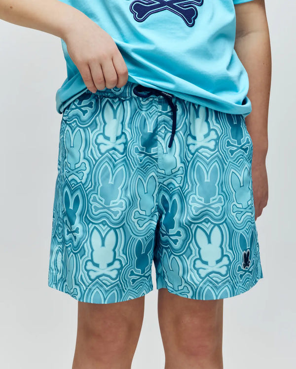 A child is shown from the waist down, wearing Psycho Bunny KIDS CHUCK ALL OVER PRINT LIGHTWEIGHT SWIM TRUNK (KB1000002) in light blue with a blue and white abstract Bunny print, paired with a matching bunny t-shirt.