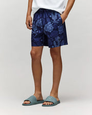 A person stands with one hand in their pocket, wearing a white shirt and Psycho Bunny’s navy KIDS ROWEN SWIM TRUNK (KB1000009), paired with teal slide sandals against a neutral background.