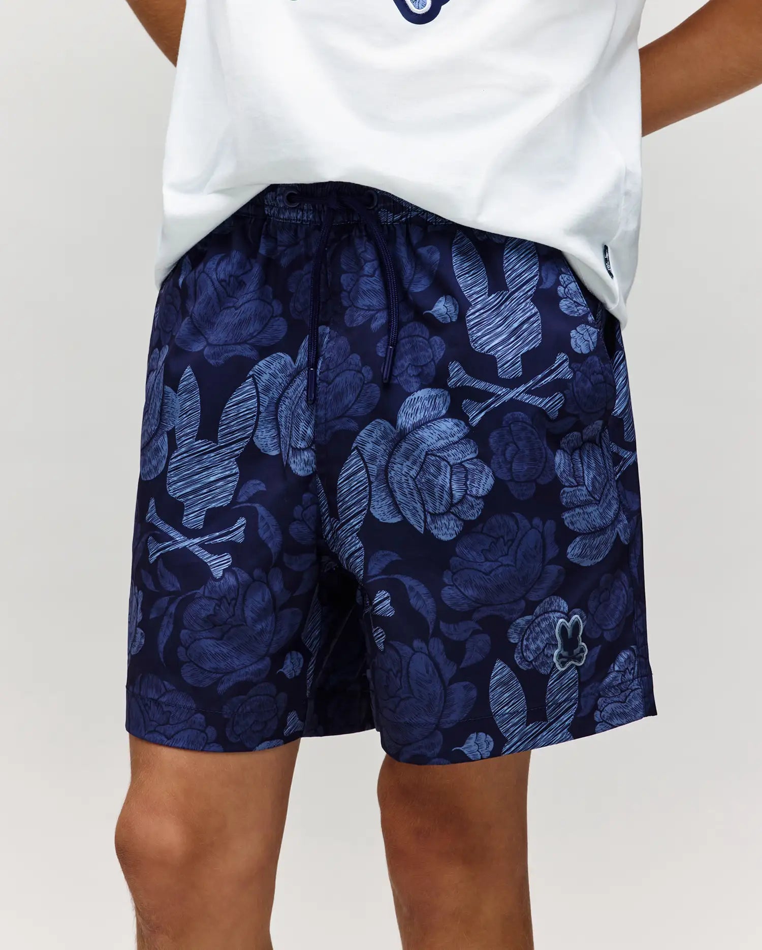 A child wears Psycho Bunny’s KIDS ROWEN SWIM TRUNK (KB1000009), featuring bold blue and light patterns, paired with a white tee. Only the lower torso and upper legs are shown, highlighting these quick-dry swim trunks.