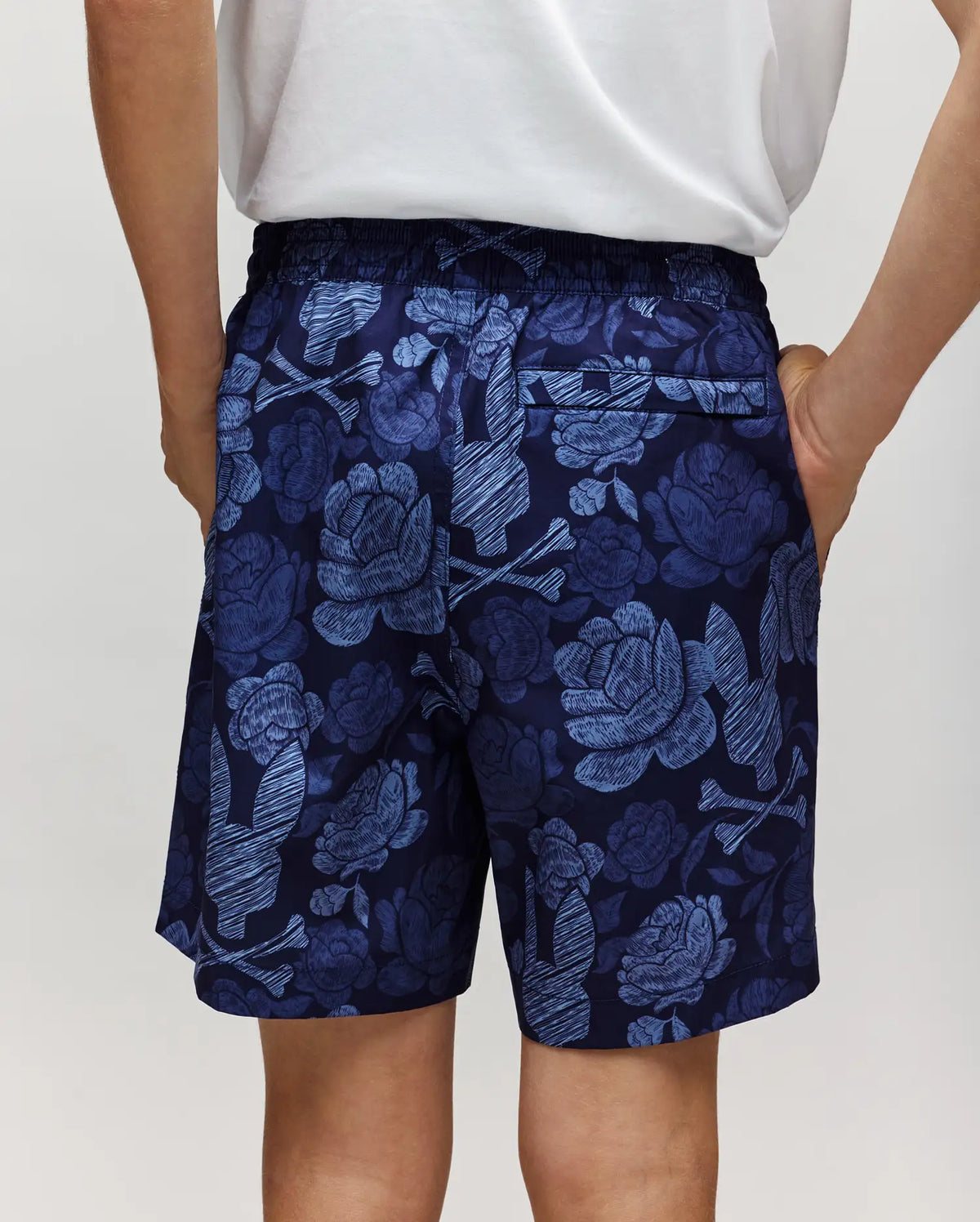 A person stands with hands in their pockets, seen from the back, wearing Psycho Bunny KIDS ROWEN SWIM TRUNK - KB1000009 in navy blue with a light blue floral pattern and a white shirt.