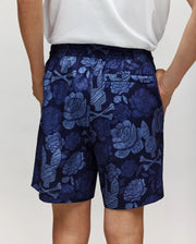 A person stands with hands in their pockets, seen from the back, wearing Psycho Bunny KIDS ROWEN SWIM TRUNK - KB1000009 in navy blue with a light blue floral pattern and a white shirt.