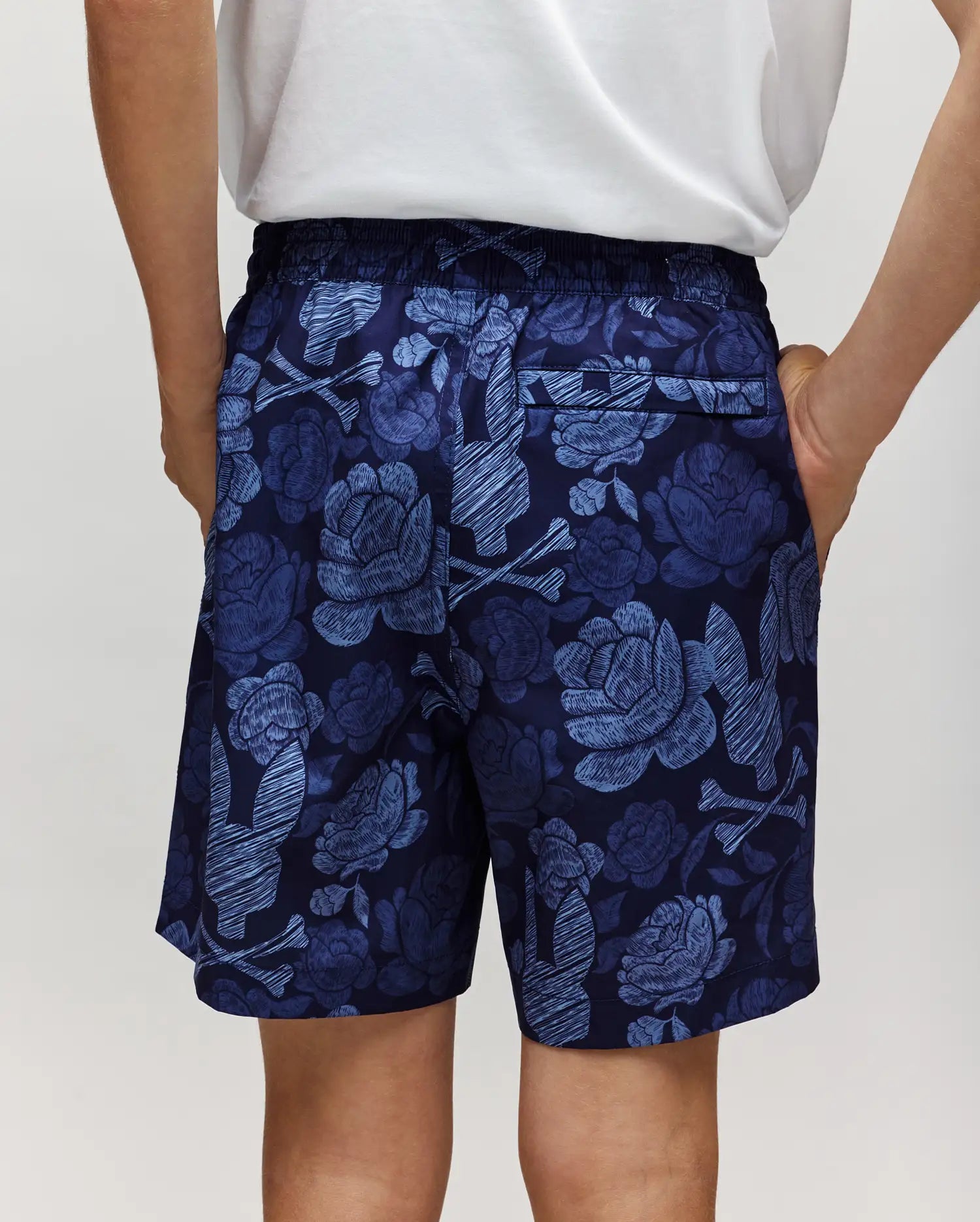 A person stands with hands in their pockets, seen from the back, wearing Psycho Bunny KIDS ROWEN SWIM TRUNK - KB1000009 in navy blue with a light blue floral pattern and a white shirt.