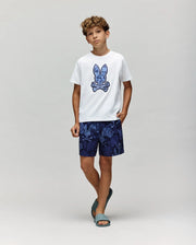 A boy with curly hair wears a white tee with a blue bunny graphic, Psycho Bunny KIDS ROWEN SWIM TRUNK - KB1000009 in dark blue floral print, and teal sandals, standing against a light gray background.