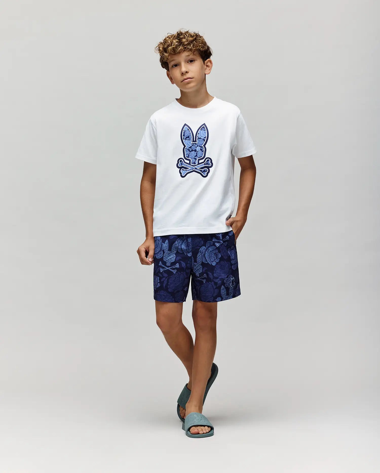 A boy with curly hair wears a white tee with a blue bunny graphic, Psycho Bunny KIDS ROWEN SWIM TRUNK - KB1000009 in dark blue floral print, and teal sandals, standing against a light gray background.