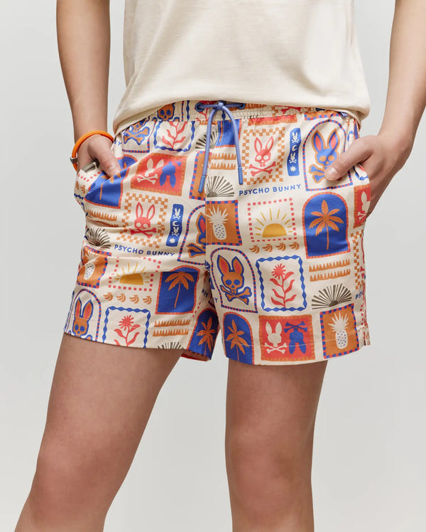 A person wears the Psycho Bunny KIDS SUNDEW SWIM TRUNK (KB1000014), featuring an allover Bunny graphic in orange, blue, and beige patchwork with palm trees, flowers, and suns. They pair it with a white top and have their hands in the pockets.